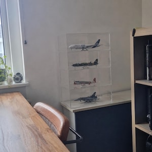 May include: A clear acrylic four-tier display case containing four distinct model airplanes. The top shelf holds a white passenger jet, the second a dark military aircraft, the third a silver propeller plane, and the bottom shelf a grey fighter jet.