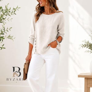May include: A long-sleeved beige blouse featuring a round neckline and a vertical row of small buttons along the side hem. The blouse is paired with white trousers and is held alongside a tan clutch bag, showcasing a neutral color palette.