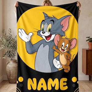May include: A rectangular fleece blanket featuring a large yellow circle on a black background. The graphic depicts the cartoon characters Tom the grey cat and Jerry the brown mouse. Below the characters, the word NAME is printed in bold yellow letters.