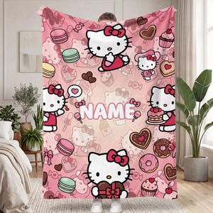 May include: A pink fleece blanket featuring a repeating pattern of Hello Kitty characters, macarons, donuts, cupcakes, and heart-shaped chocolates. The center of the blanket displays the word NAME in bold white letters with a pink outline.