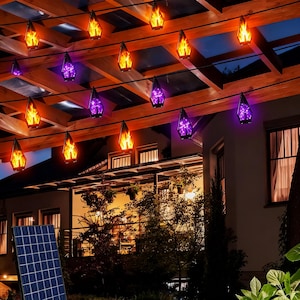 May include: A string of outdoor solar-powered lantern lights hanging from a wooden pergola. The lanterns feature alternating orange flame-effect lights and solid purple lights, suspended on black wires above a solar panel in the foreground.