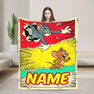 May include: A rectangular throw blanket featuring a comic book style graphic of Tom the cat bursting through a red background and Jerry the mouse running across a yellow background, with the word NAME printed in large yellow letters at the bottom.