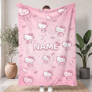May include: A light pink fleece blanket featuring a repeating pattern of Hello Kitty characters in various poses, interspersed with small bows and dots. The word NAME is printed in large, white, rounded capital letters in the center.
