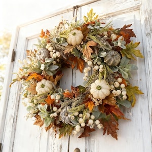 May include: A decorative wreath featuring white pumpkins, grey pumpkins, and autumn-colored maple leaves in shades of orange, yellow, and brown. The arrangement includes clusters of small white berries and green foliage accents.