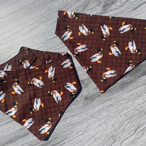 May include: Two triangular pet bandanas featuring a brown plaid background with a repeating pattern of mallard ducks. Each bandana is made of fabric and includes a visible metal snap fastener for securing the accessory around a pet's neck.