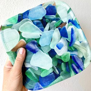 May include: A square, light blue, green, and brown fabric coaster with a pattern of sea glass. The coaster is made of a soft, textured fabric.