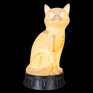 May include: A glowing, translucent cat-shaped lamp with a visible internal skeletal structure. The cat sits upright on a circular black base featuring a decorative arched pattern. The light emits a warm, soft yellow hue throughout the body.