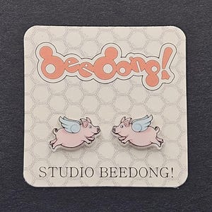 May include: A pair of stud earrings featuring pink flying pigs with light blue wings. The earrings are attached to a square display card with a white honeycomb pattern, the text "beeDong!" in large peach letters, and "STUDIO BEEDONG!" at the bottom.