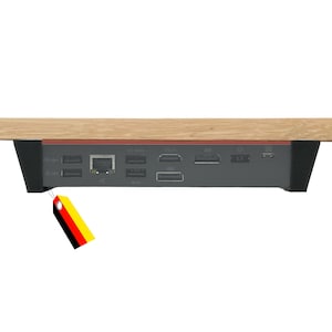 Könnte beinhalten: Eine dunkelgraue elektronische Dockingstation mit verschiedenen Anschlüssen wie USB, Ethernet, HDMI und DisplayPort, einer hellen Holzoberfläche und schwarzen Seitenstützen, ergänzt durch ein kleines Etikett mit schwarzen, roten und goldenen Streifen.