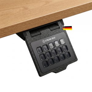 Könnte beinhalten: Eine schwarze Stream Deck Steuerungskonsole mit fünfzehn quadratischen Tasten, die unter einer Holzoberfläche montiert ist. Das Gerät verfügt über einen strukturierten Rahmen, eine Scharnierhalterung und den weißen Schriftzug STREAM DECK.