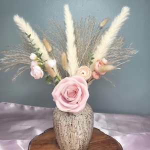 May include: A floral arrangement featuring a large central pink rose, smaller peach-colored blossoms, and tall white pampas grass plumes. The bouquet is displayed in a speckled, light brown ceramic vase with a rounded, tapered shape.