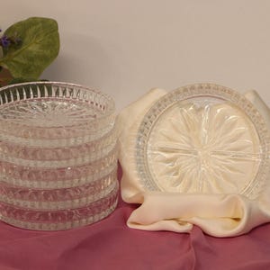 May include: A set of five clear glass coasters with a circular shape and a pressed starburst pattern in the center. The coasters feature a vertical ribbed edge design and are stacked together, with one coaster displayed upright in a cream fabric fold.