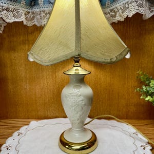 May include: A table lamp with a cream-colored ceramic base featuring a raised grape cluster design and a polished gold-tone metal base. The lamp is topped with a flared, textured gold fabric shade that emits a warm, soft glow when illuminated.