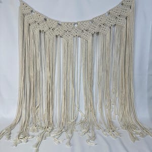 May include: A cream-colored macrame wall hanging featuring an intricate knotted geometric pattern along the top edge, transitioning into long, loose, vertical fringe strands that hang down to create a textured, layered appearance.