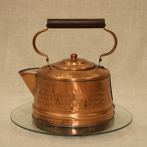 May include: A hammered copper tea kettle with a polished finish, featuring a rounded body, a short spout, and a dark brown wooden handle attached to a curved metal frame that arches over the top of the lid and the main body of the kettle.