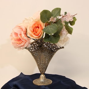 May include: A metallic, antique-finish vase with an intricate, openwork filigree rim and a tapered, embossed body. The vase is filled with a bouquet of artificial flowers, including large peach-colored roses and green eucalyptus-style leaves.