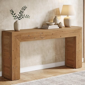 May include: A rectangular wooden console table with a natural grain finish. On top sits a white table lamp, a stack of two books, a small bowl, a small decorative vase, and a white ceramic vase containing a green leafy branch.