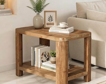 May include: A rectangular wooden side table with four thick legs and a lower shelf. The top surface holds a light-coloured vase with a green plant, a framed print, and a stack of books with a white coffee cup. The lower shelf holds more books.