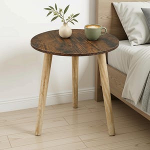 May include: A round side table with a dark brown wood grain top and three light-coloured tapered wooden legs. A small cream-coloured vase containing a green leafy branch and a sage green ceramic mug sit on the surface of the table.
