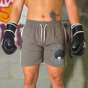 Könnte beinhalten: Graue Sportshorts mit weißem Kordelzug am Bund und weißen Seitenstreifen. Die Shorts zeigen eine Grafik einer Figur mit großem Afro und den in Blau gedruckten Schriftzug "AFRO LUFFY" auf dem unteren Bereich des rechten Hosenbeins.