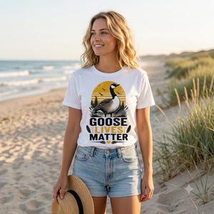Może przedstawiać: Biały t-shirt z krótkim rękawem z grafiką przedstawiającą gęś na tle żółtego słońca i drzew. Napis GOOSE LIVES MATTER jest nadrukowany w kolorze czarnym i żółtym pod gęsią, wraz z małymi ikonami piór i małym sercem.