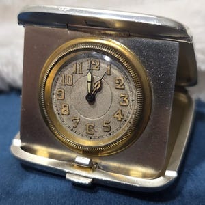 May include: A silver-toned metal travel clock housed in a hinged case. The clock face is circular with a gold-colored bezel, featuring black Arabic numerals from 1 to 12 and two black hands. The case has a smooth, metallic finish.
