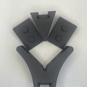 Könnte beinhalten: Ein Set aus vier dunkelgrauen 3D-gedruckten Komponenten, bestehend aus einer großen V-förmigen Basis mit geschwungenen Seiten, einem zentralen dreieckigen Keilstück und zwei rechteckigen Seitenplatten mit jeweils zwei erhabenen Kreisdetails.