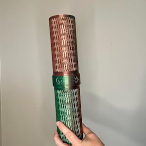 May include: A cylindrical container featuring a metallic copper top section and a metallic green bottom section, both with a diamond-shaped textured pattern. A central band separates the two halves, displaying a small skull icon and a sun icon.