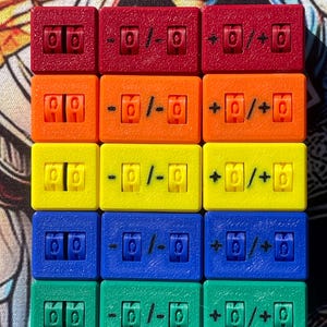 May include: A set of fifteen rectangular plastic counters arranged in a five-by-three grid. Each row features a distinct color: red, orange, yellow, blue, and green. The counters display numerical digits and mathematical symbols including plus and minus signs.