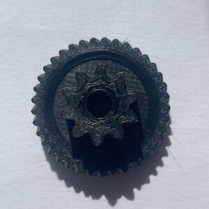 Könnte beinhalten: Ein schwarzes Kunststoff-Zahnrad mit einem größeren äußeren Ring mit zahlreichen kleinen Zähnen und einem kleineren, erhöhten inneren Zahnrad mit zehn ausgeprägten Zähnen, das um eine kreisförmige Öffnung in der Mitte angeordnet ist.
