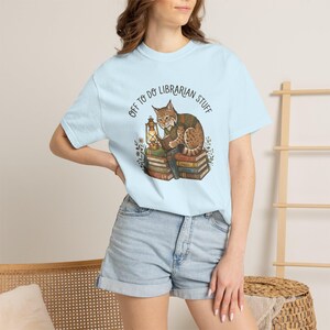 May include: A light blue short-sleeve t-shirt featuring a graphic of a brown lynx wearing a green vest, sitting on a stack of books while reading and holding a lantern. The text above the illustration reads, "OFF TO DO LIBRARIAN STUFF."