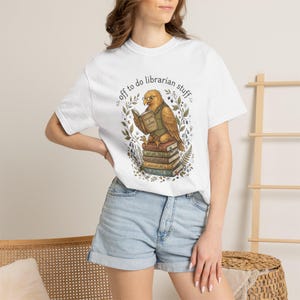 Puede incluir: Una camiseta blanca de manga corta con un gráfico de un búho con gafas y chaleco, leyendo un libro titulado Folklore & Legends. Debajo del búho hay una pila de libros etiquetados como Stories, Nature Tales, Wilderness, Adventures, Plants.