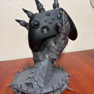 May include: A matte gray 3D-printed controller stand shaped like a clawed, armored hand emerging from a circular, spiked base. The hand is gripping a black wireless gaming controller with a central Xbox logo and multicolored action buttons.