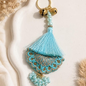 May include: A light blue keychain featuring a gold metal ring, two small gold bells, and a strand of light blue beads. A light blue tassel sits above a semi-circular embroidered fabric piece with gold thread accents and a cluster of light blue beads.