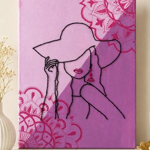 May include: A rectangular canvas wall art piece featuring a split-tone light and dark purple background with magenta mandala patterns. A black thread outline depicts a figure wearing a wide-brimmed hat, with embroidered red lips and a red earring.