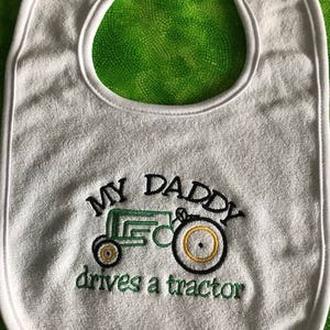 May include: White baby bib with green and black embroidery of a tractor and the text "My Daddy drives a tractor".