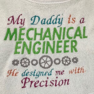 Puede incluir: Babero blanco para beb&eacute; con texto bordado de colores que dice "Mi pap&aacute; es un INGENIERO MEC&Aacute;NICO Me dise&ntilde;&oacute; con Precisi&oacute;n". El texto est&aacute; rodeado de peque&ntilde;os engranajes grises.