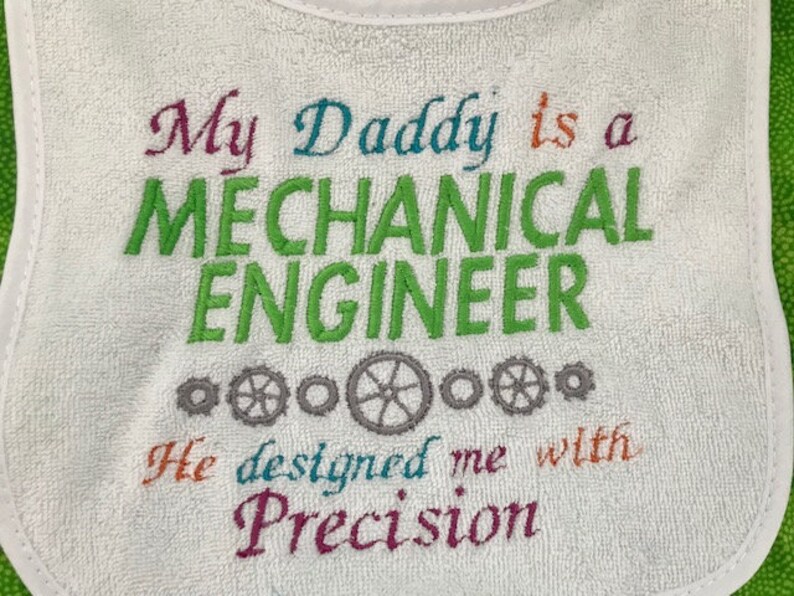 Puede incluir: Babero blanco para beb&eacute; con texto bordado de colores que dice "Mi pap&aacute; es un INGENIERO MEC&Aacute;NICO Me dise&ntilde;&oacute; con Precisi&oacute;n". El texto est&aacute; rodeado de peque&ntilde;os engranajes grises.
