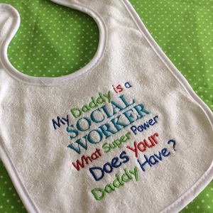 May include: White baby bib with embroidered text that reads "My Daddy is a Social Worker What Super Power Does Your Daddy Have?"
