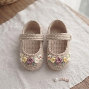 May include: A pair of beige crochet baby shoes featuring a single strap across the top. The front of each shoe is decorated with embroidered flowers in shades of yellow, white, and mauve, accented with small green leaves and a single white pearl.