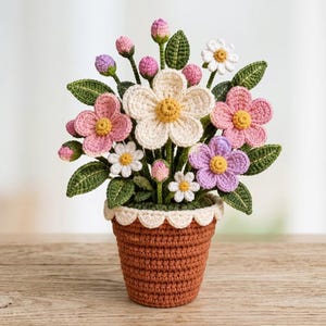 Puede incluir: Un arreglo floral de ganchillo en una maceta marrón con un borde festoneado color crema. El ramo presenta flores de cinco pétalos blancas, rosas y moradas con centros amarillos, junto con pequeños capullos rosas y hojas verdes texturizadas.