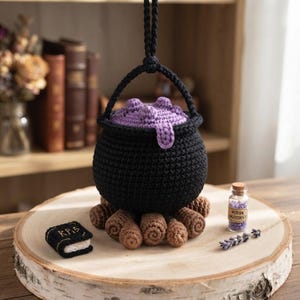 May include: A black crocheted cauldron with a purple bubbling liquid top, suspended by a black handle. It sits atop brown crocheted logs. Beside it is a small black book with the text "K P I S" and a glass vial labelled "POTION INGREDIENT".