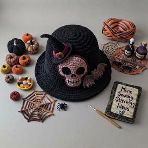 Op de afbeelding: Een zwarte gehaakte bucket hat met een roze schedelapplicatie, een kleine heksenhoed en een vleermuis. Omringende items zijn gehaakte pompoenen, een spinnenweb, een mandje met snoepjes en een boek met de titel More Spooky Stitching Ideas.