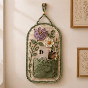 Puede incluir: Un organizador de pared de ganchillo con base color crema y un bolsillo verde salvia. La pieza presenta un tulipán morado grande y una margarita blanca con centro amarillo. Un teléfono inteligente y unas gafas están dentro del bolsillo.