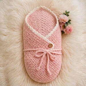 May include: A soft pink crochet baby swaddle blanket featuring a white scalloped edge trim, a single wooden button closure, and a matching pink yarn tie. The swaddle is oval-shaped with a textured knit pattern and is accented by small pink flowers.