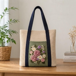 May include: A beige crochet tote bag featuring dark navy blue handles and a central sage green panel. The panel is decorated with a three-dimensional floral arrangement consisting of crocheted pink roses, white daisies, and small purple buds.