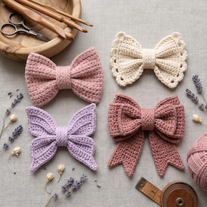 May include: Four crocheted bows in dusty pink, cream, lavender, and mauve, arranged with wooden crochet hooks, metal scissors, dried lavender sprigs, a ball of yarn, and a wooden measuring tape on a neutral linen fabric background.