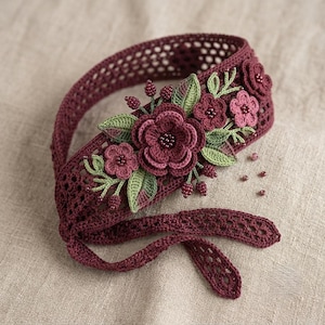 May include: A deep burgundy crochet headband featuring an intricate openwork pattern. The center is adorned with layered dark red and pink crocheted flowers, sage green leaves, and small dark red beaded accents scattered across the surface.