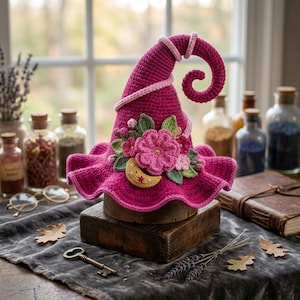 May include: A vibrant magenta crocheted hat with a curled tip, featuring a light pink spiral trim. The hat is decorated with a cluster of crocheted flowers in shades of pink and green, along with a small yellow crescent moon with red star accents.