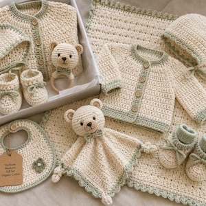 Puede incluir: Un conjunto de regalo para bebé tejido a ganchillo en hilo color crema y verde salvia. Incluye dos cárdigans con botones de madera, dos gorritos, dos pares de patucos, un babero con la etiqueta Heirloom Newborn Gift Set Organic Cotton, un sonajero de oso y una manta.